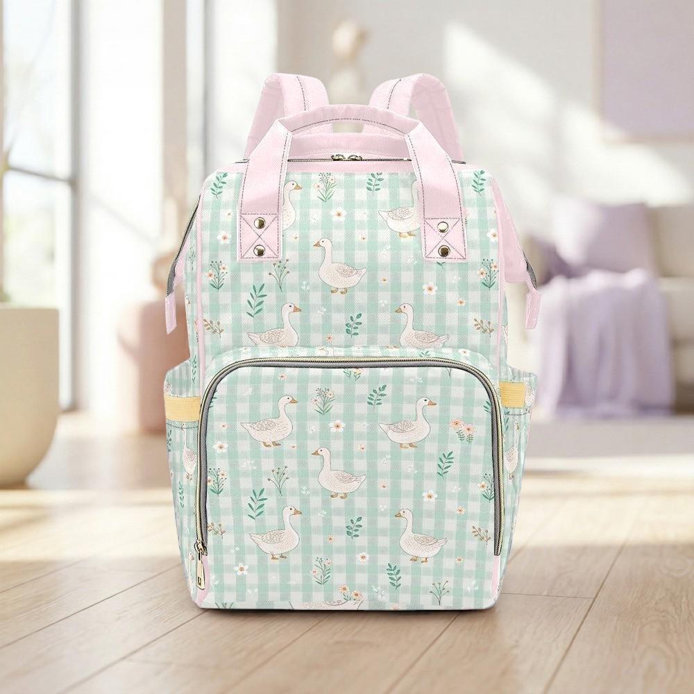 A pink and green gingham-patterned backpack with a floral design and white ducks printed on it, placed on a wooden floor against a white background.