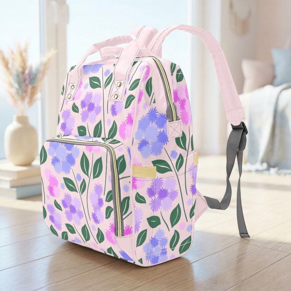 A pink backpack with a floral pattern is sitting on a wooden floor in a room with a window and a vase of flowers in the background.