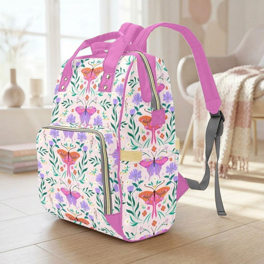 A colorful floral backpack with a pink strap is sitting on a wooden floor in a room with a window and a chair visible in the background.