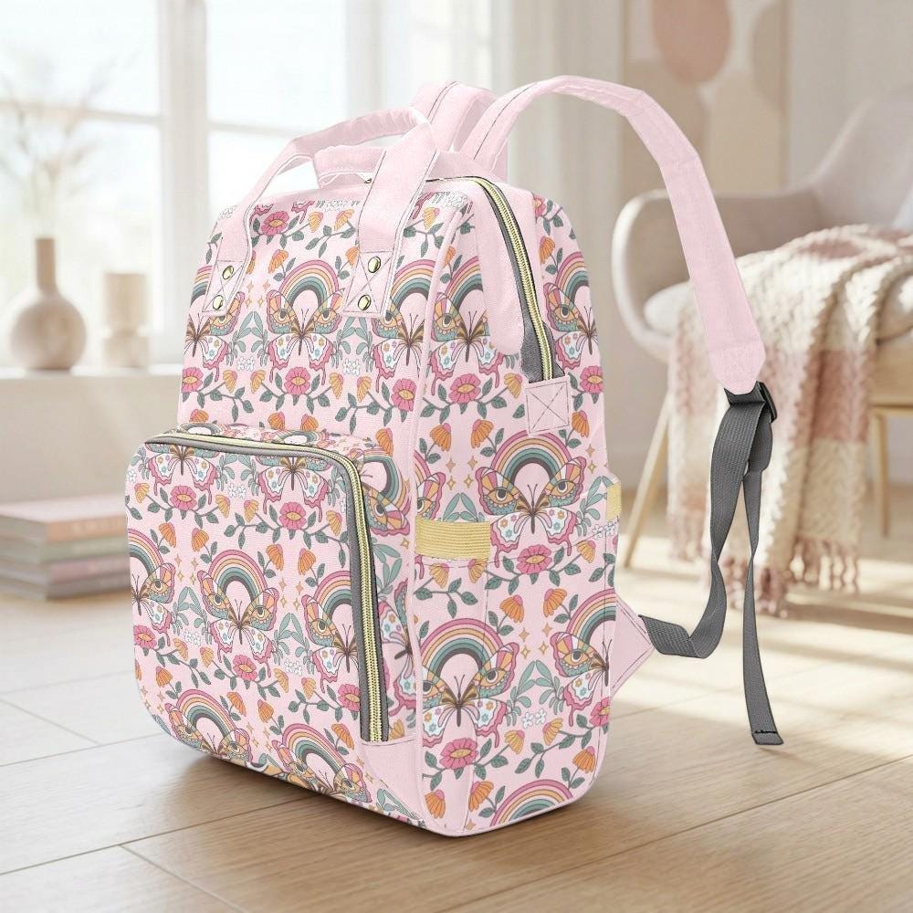A pink backpack with a floral pattern and a rainbow design sits on a wooden floor in a room with a window and a chair.