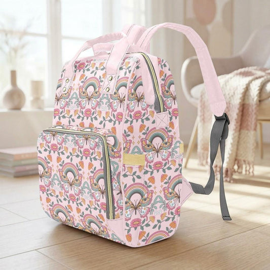 A pink backpack with a floral pattern and a rainbow design sits on a wooden floor in a room with a window and a chair.