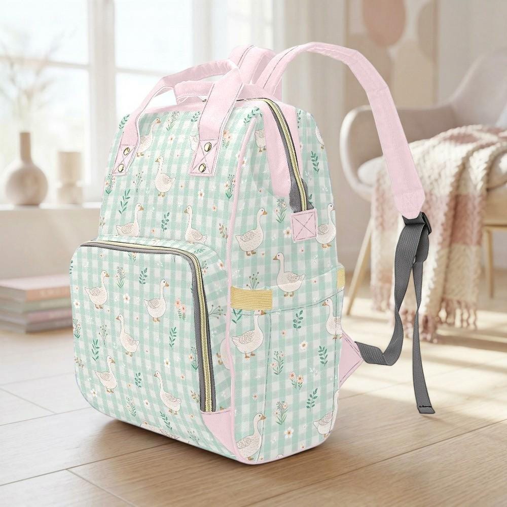 A pink and green gingham-patterned backpack with a floral design and ducks printed on it, sitting on a wooden floor in a room with a window and a chair visible in the background.